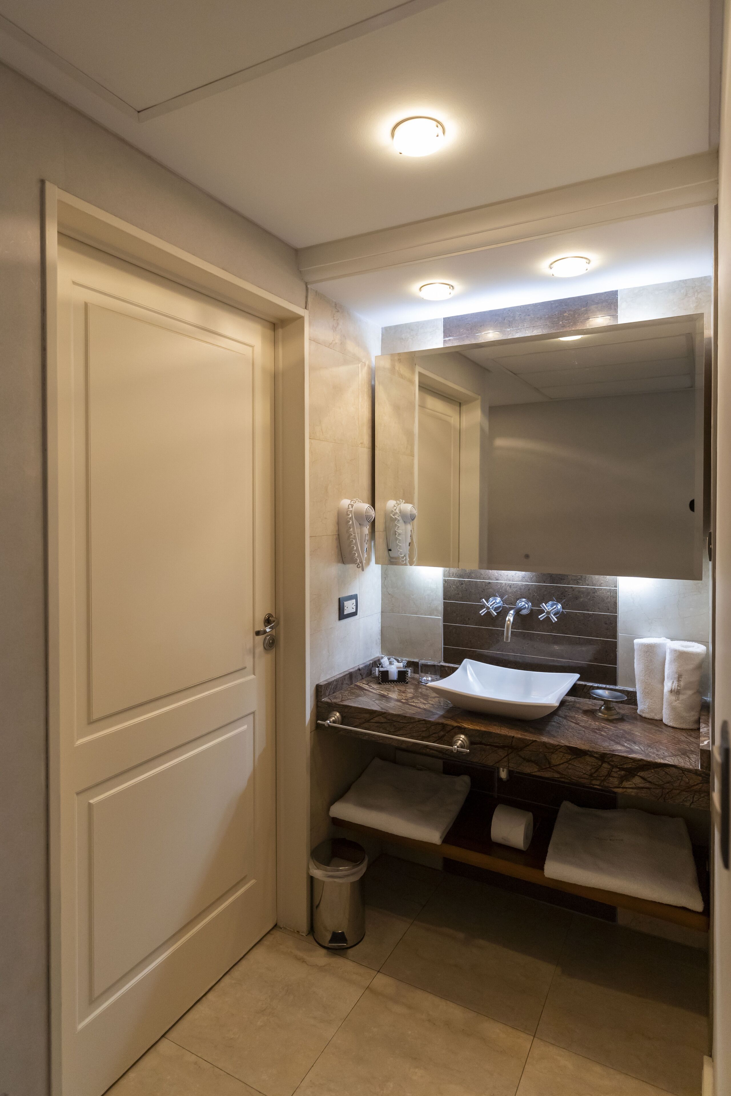 Un baño con una puerta blanca, tocador con azulejos de mármol, lavamanos y espejo.