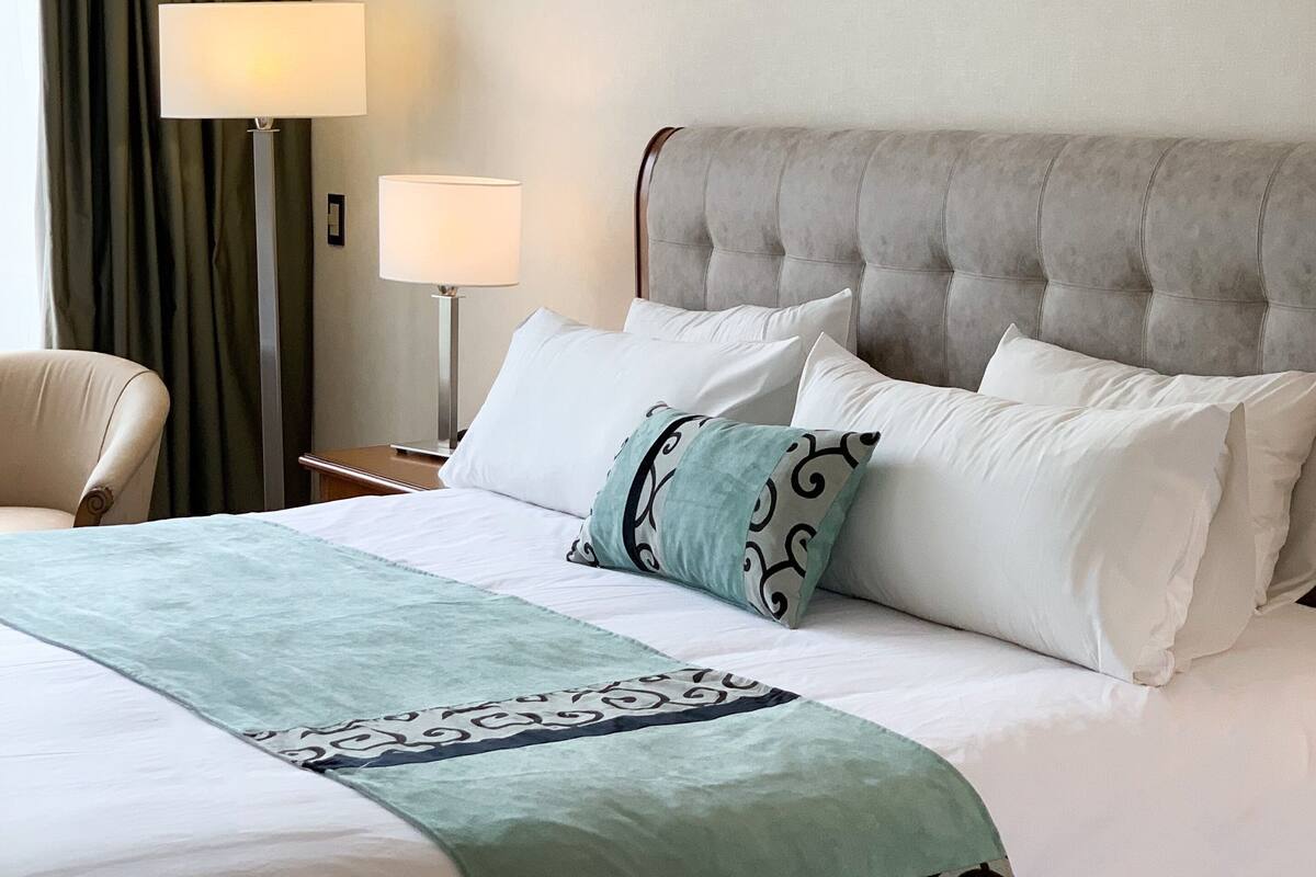 A hotel room with a large bed, a chair, a nightstand with a lamp, and a view of the outside through a window with curtains.