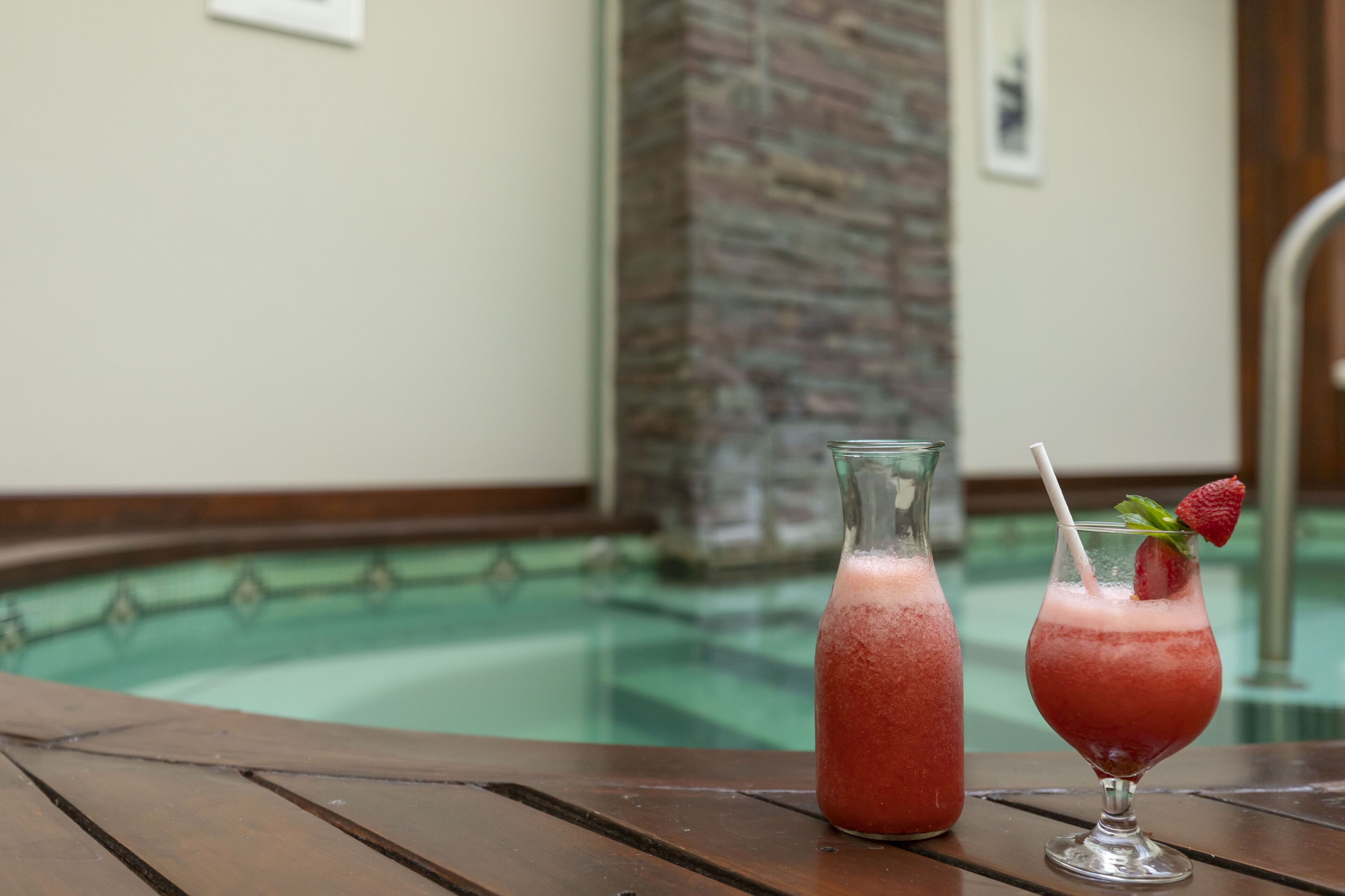 Une scène au bord de la piscine avec une table en bois, un pichet en verre et un verre décoré d’une fraise.