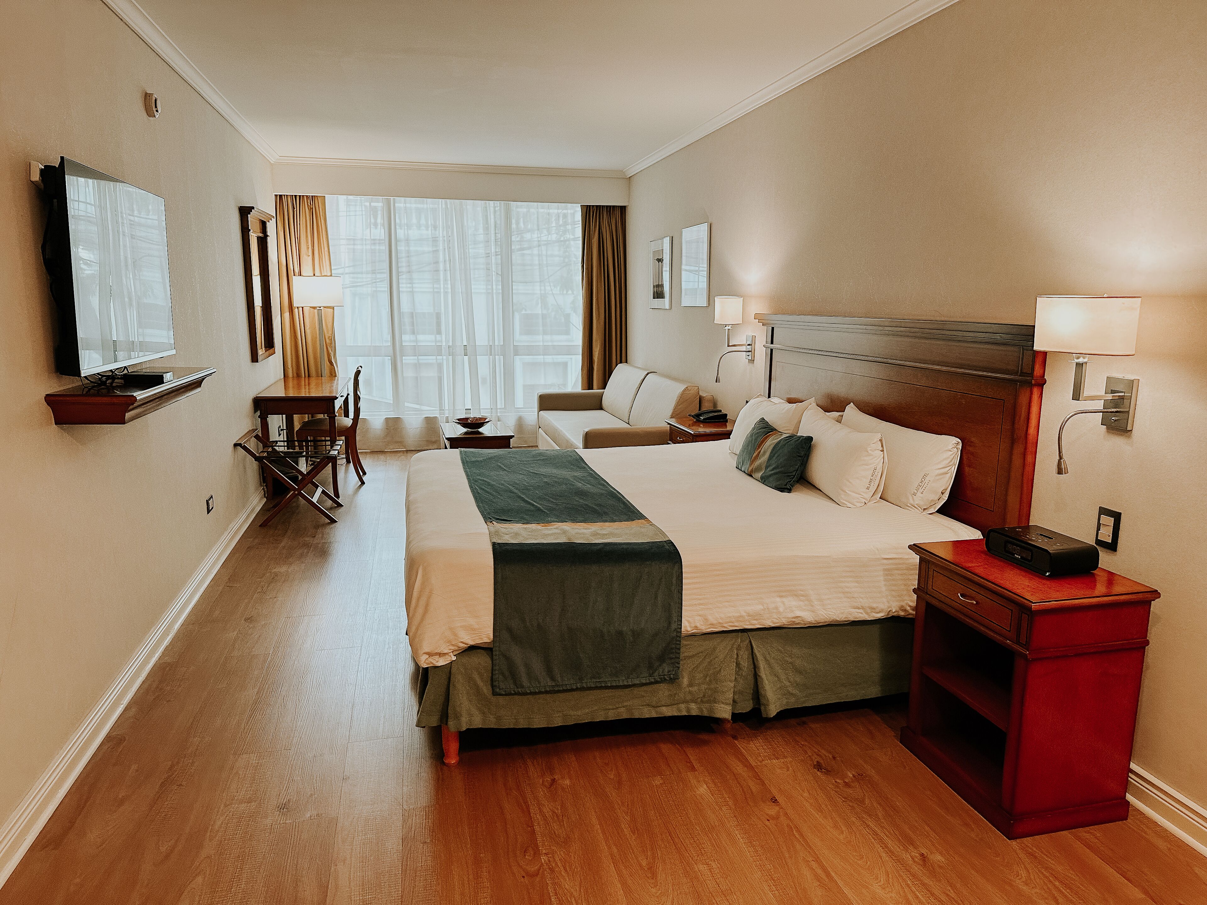 A hotel room with a large bed, a desk, a chair, a TV, and a window with curtains.