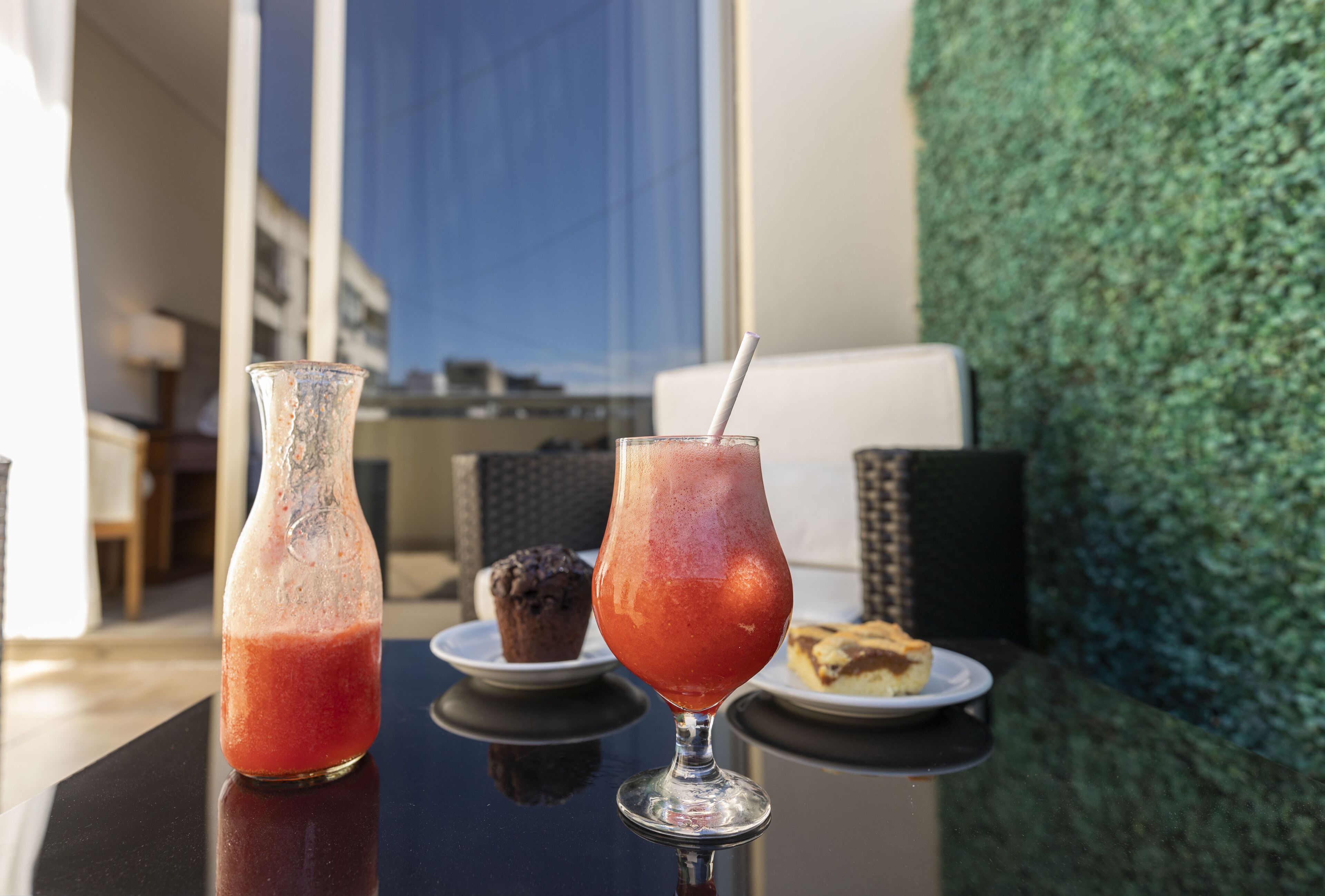 A table with a glass of red drink, a glass of iced drink, and a muffin on a plate.