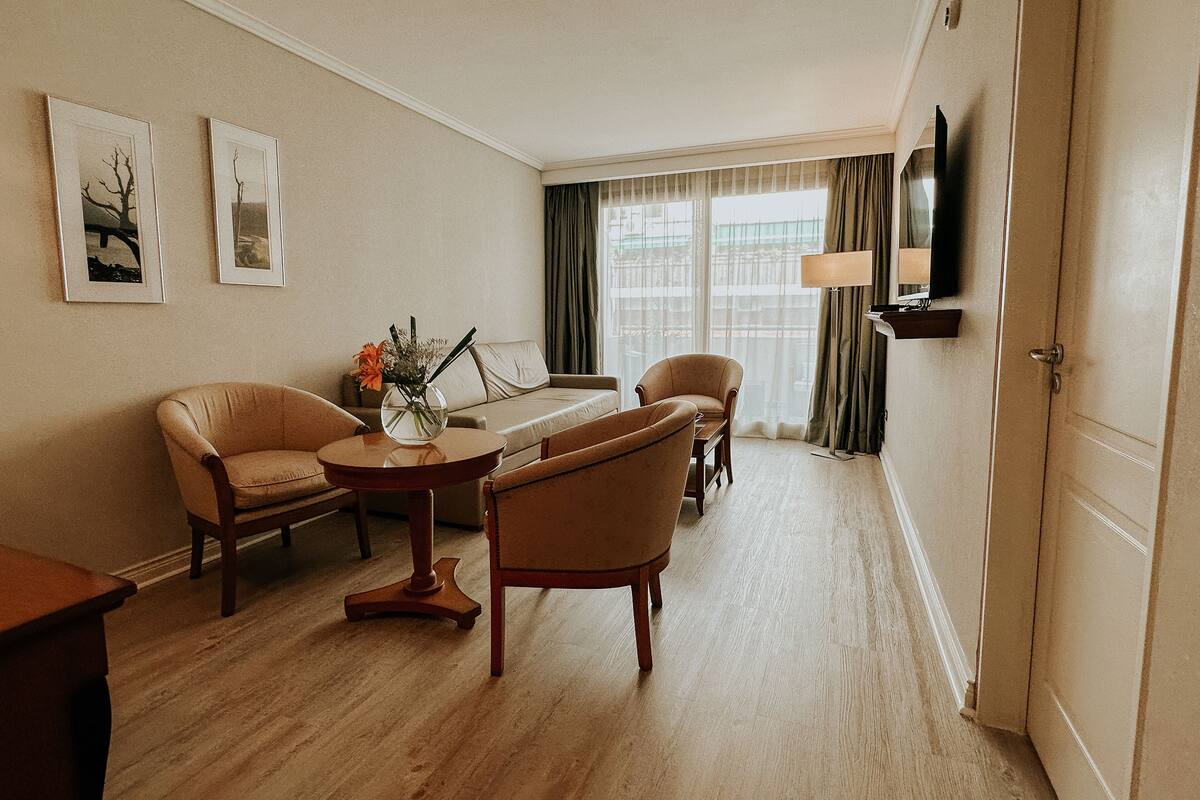 A hotel room with a wooden floor, beige walls, a sofa, two armchairs, a small round table with a vase, a flat-screen TV mounted on the wall, and a window with curtains.