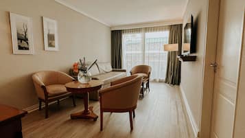 Habitación de hotel con piso de madera, paredes color beige, un sofá, dos sillones, una pequeña mesa redonda con un florero, un televisor de pantalla plana montado en la pared y una ventana con cortinas.