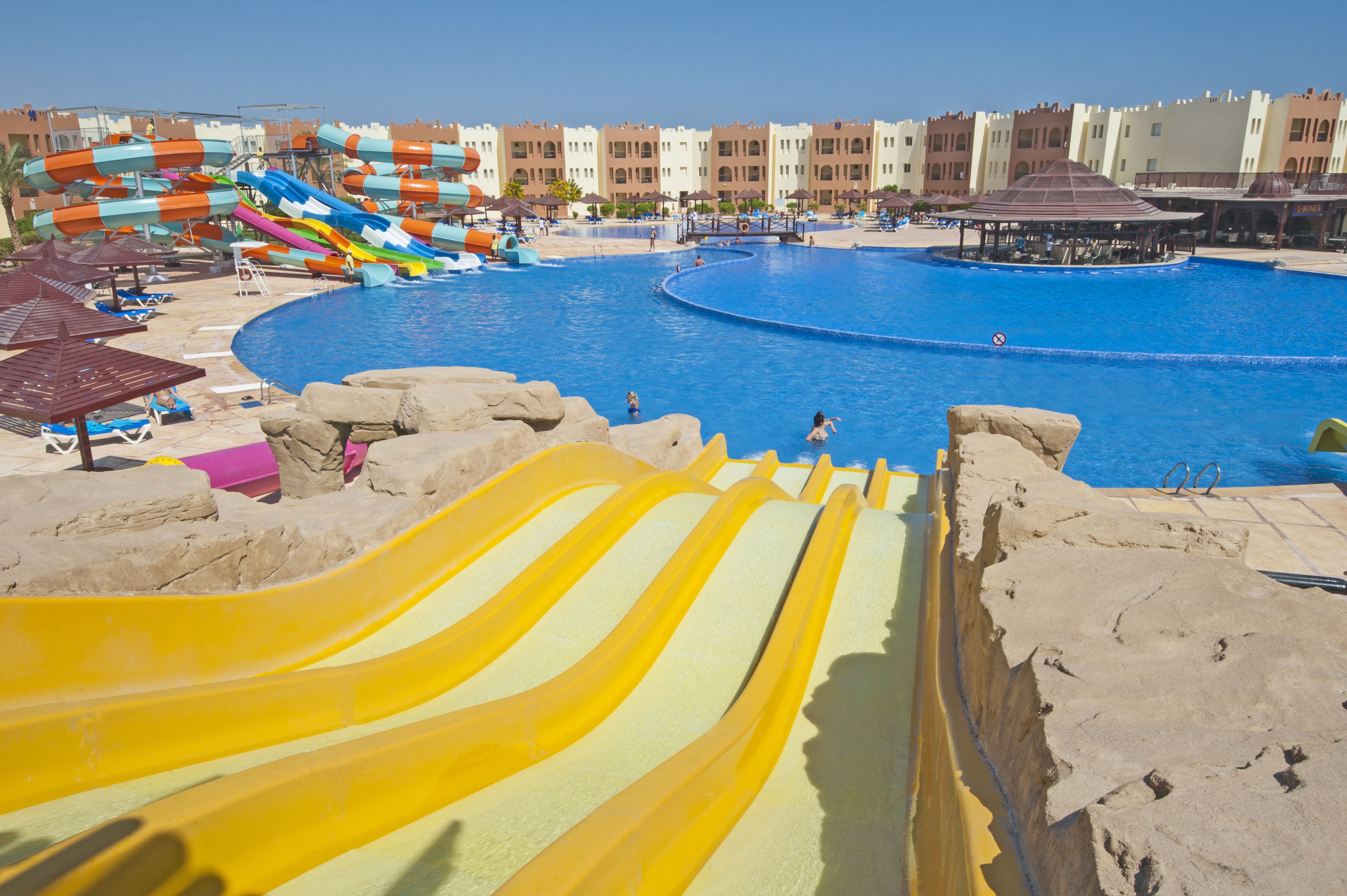 A large swimming pool with multiple slides, a sandy beach area, and a row of buildings in the background.