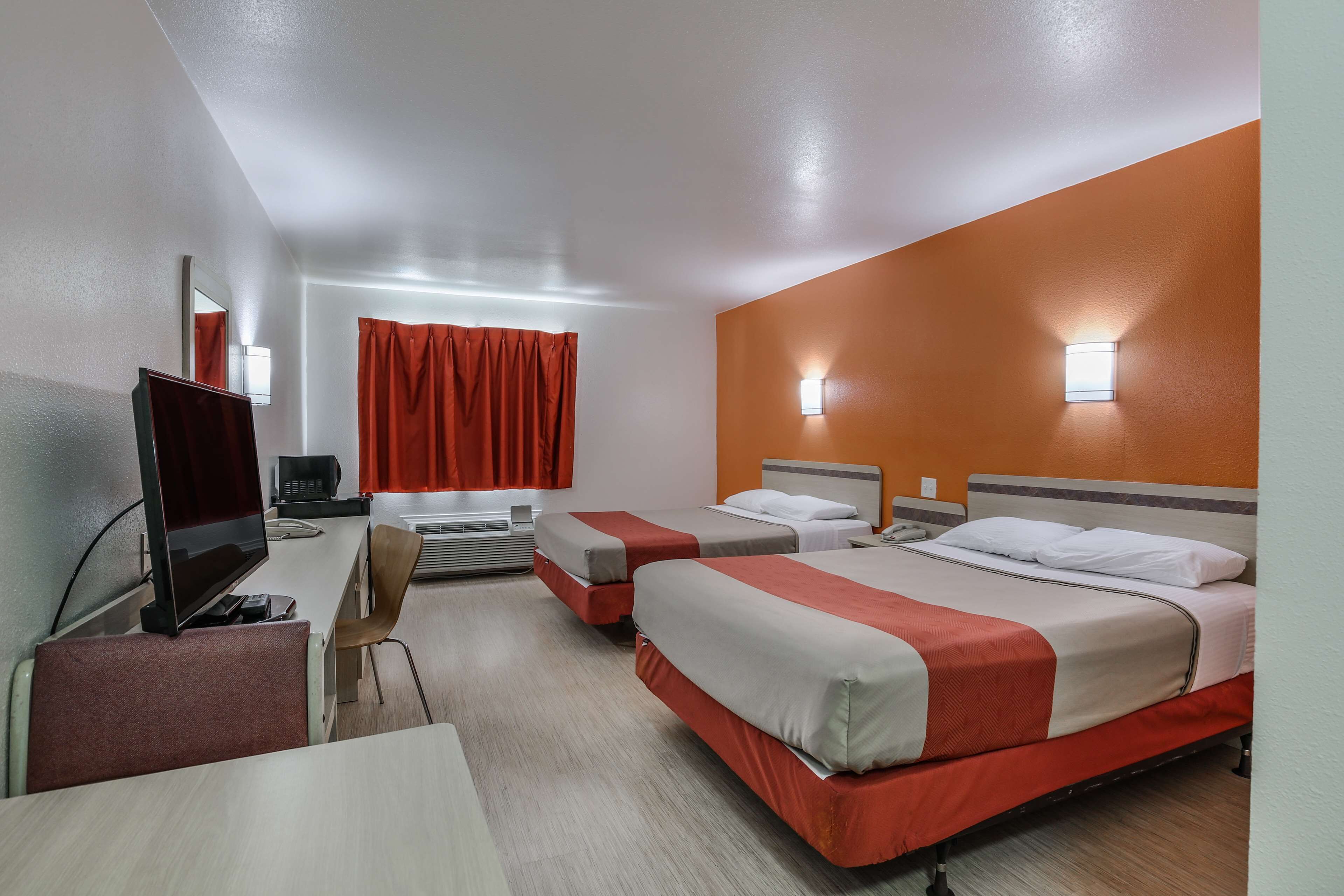 Habitación de hotel con dos camas, un escritorio con computadora, una silla y una ventana con cortinas rojas.