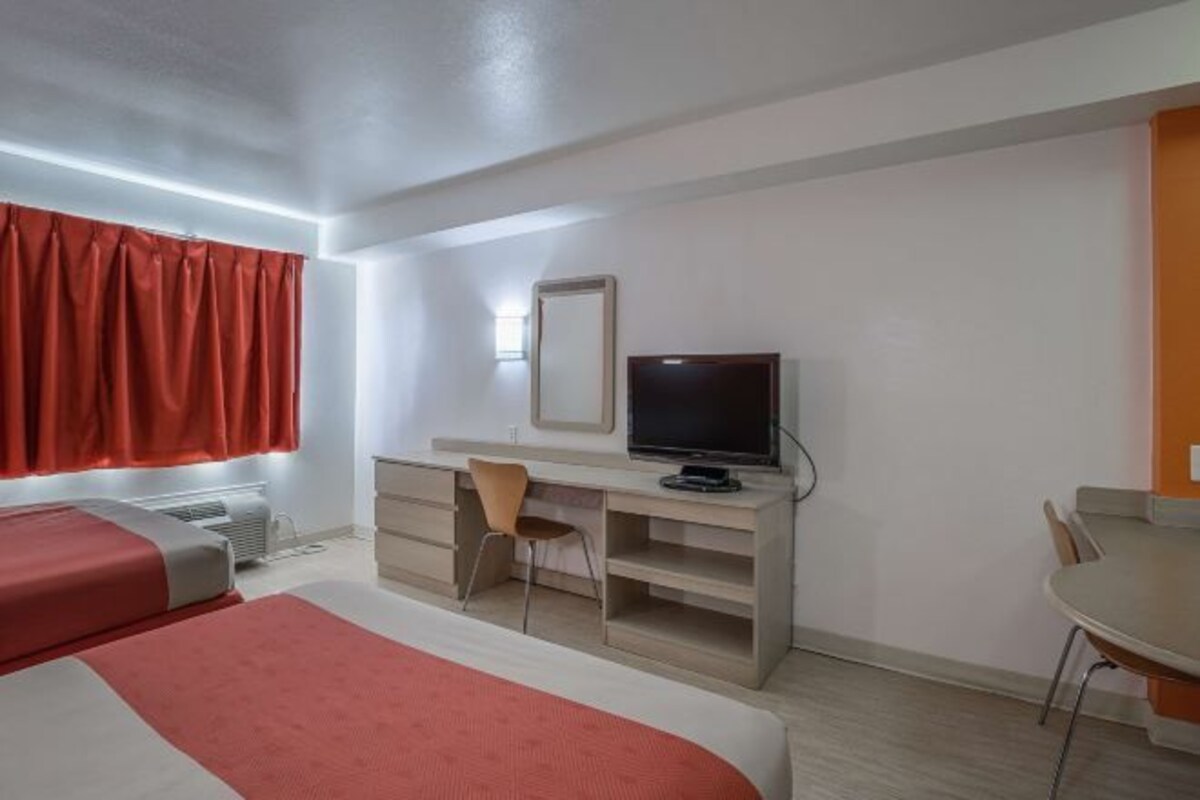 A hotel room with a bed, a desk, a chair, a TV, and a window with red curtains.
