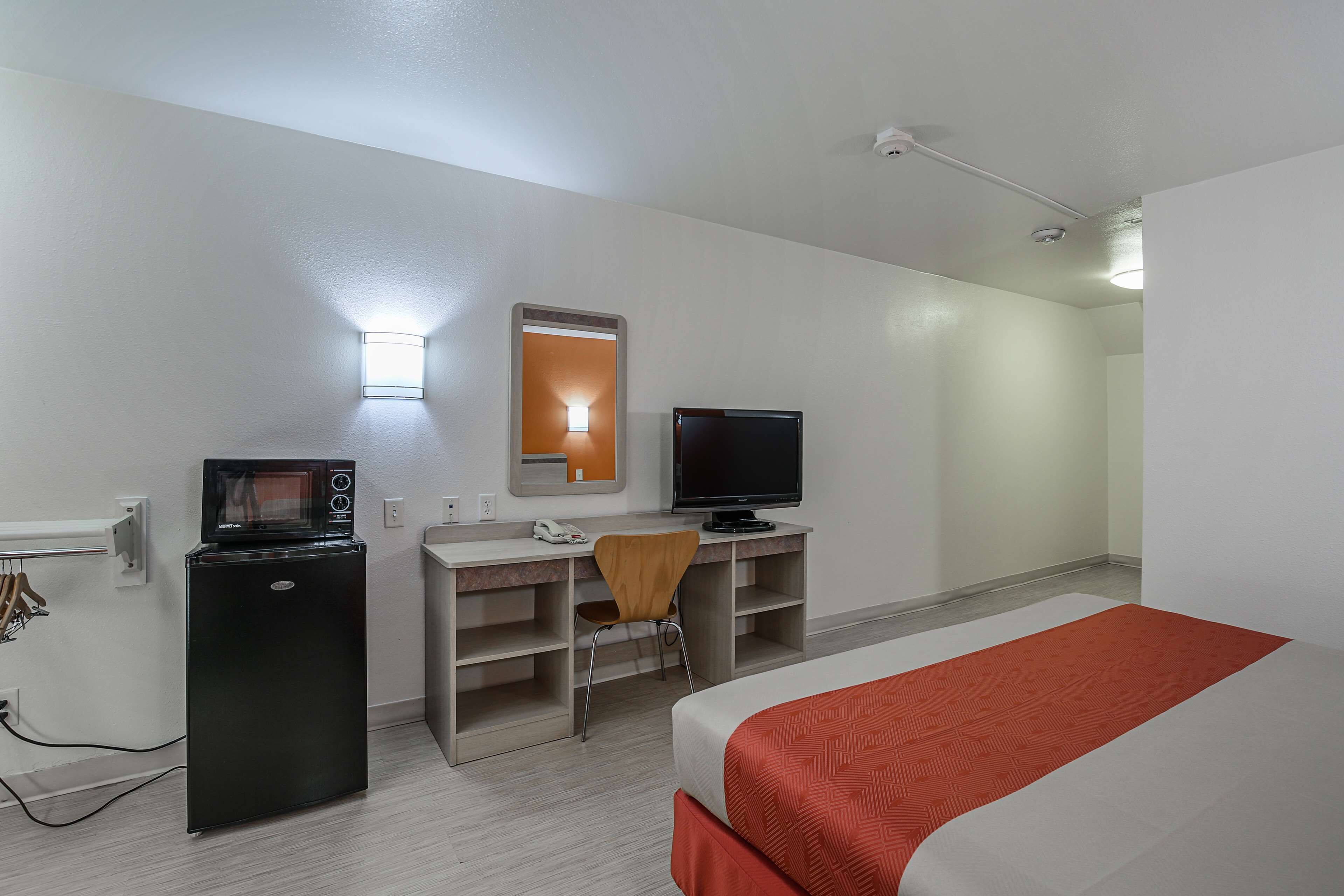 A hotel room with a bed, a desk with a computer and a microwave, a chair, and a small window.