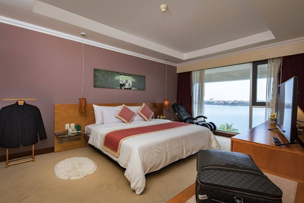 A hotel room with a bed, a desk with a TV, a chair, a coat rack, and a view of the water.