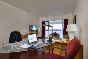 Habitación de hotel con escritorio de vidrio, una computadora portátil, televisor, sofá y vista a un cuerpo de agua.