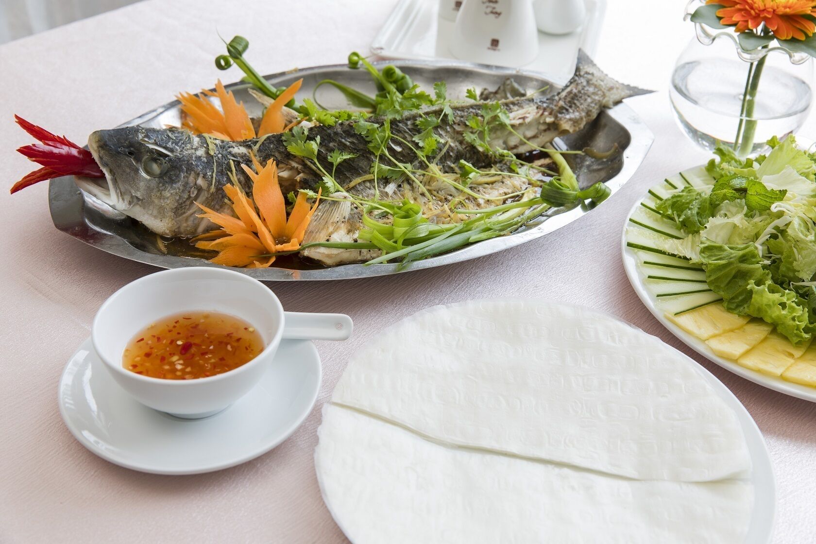 A table with a fish dish, a bowl of dipping sauce, and a plate of sliced vegetables.