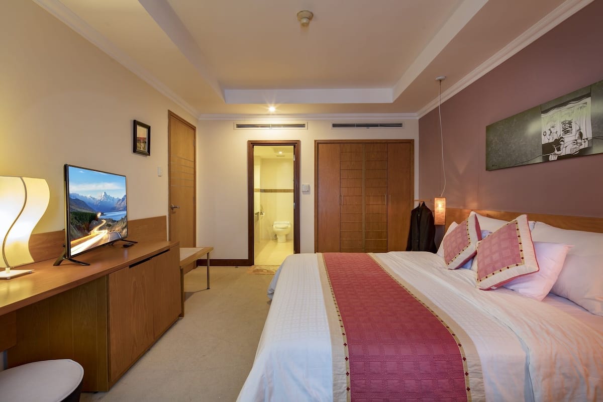 A hotel room with a bed, bedside table, lamp, TV, and a painting on the wall.