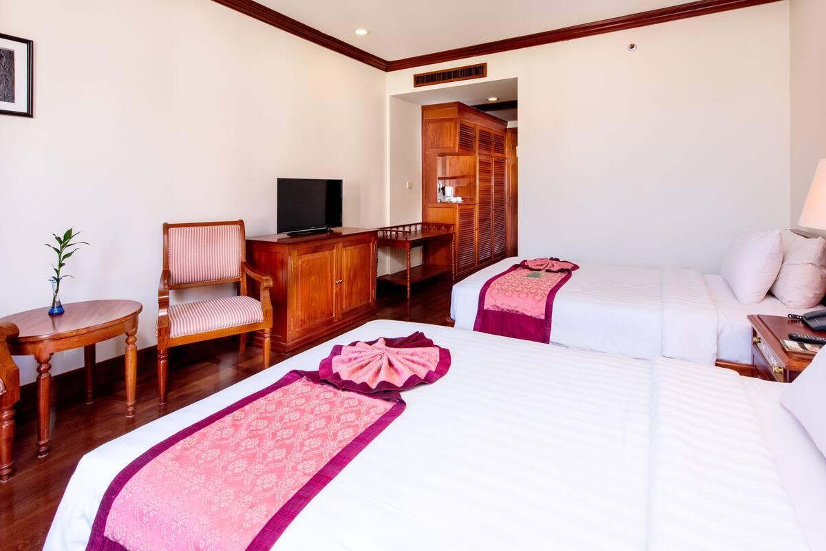 A hotel room with two beds, a desk with a television, a chair, a small table with a plant, and a wardrobe.