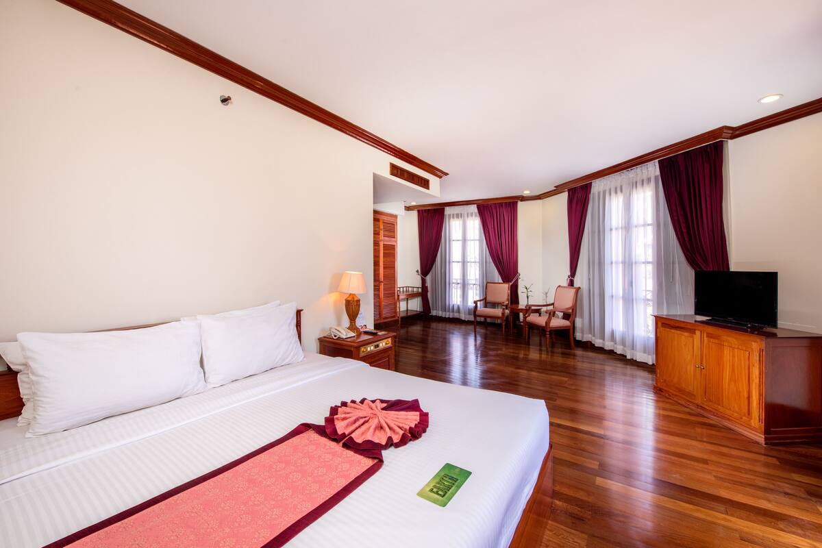 A hotel room with a large bed, wooden flooring, a TV, a desk with a lamp, and a seating area with chairs.
