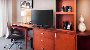 Quarto de hotel com escrivaninha de madeira, televisão de tela plana, abajur, cadeira e cafeteira.