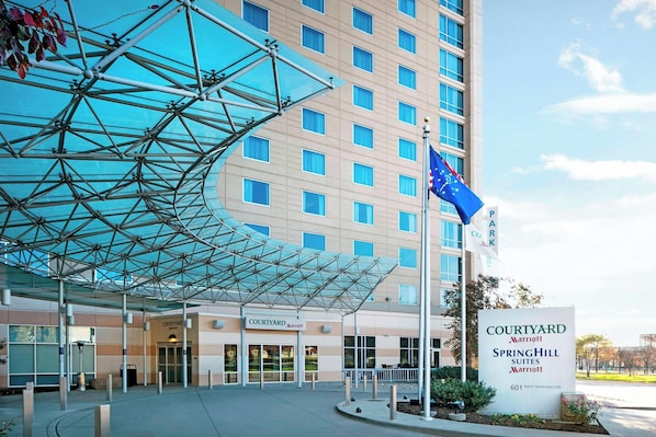 Entrada de un hotel Courtyard Marriott con un toldo de vidrio, una bandera y un letrero que indica SpringHill Suites.