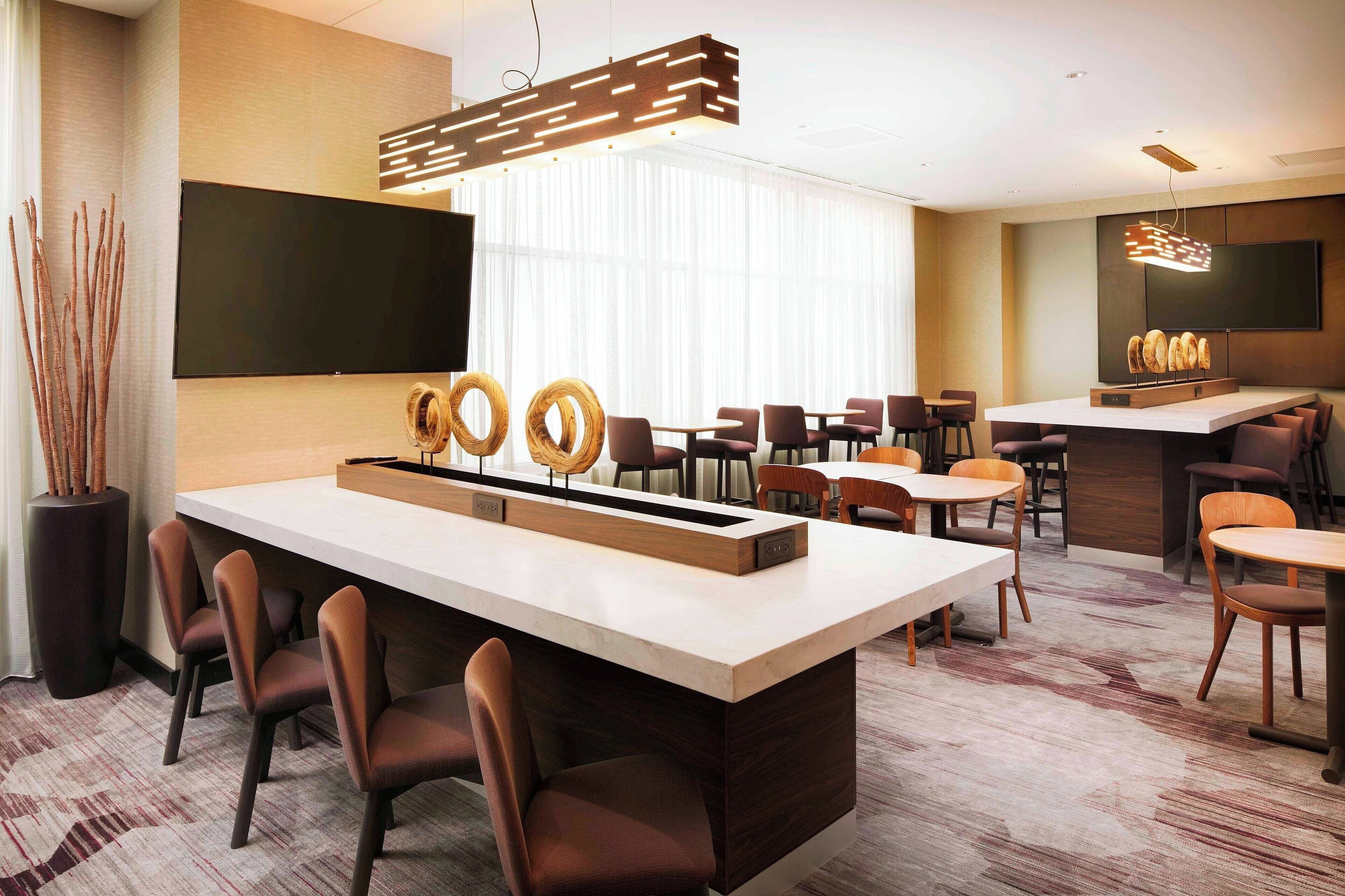 A modern hotel lobby with a central bar area, seating arrangements, and a large flat-screen TV mounted on the wall.