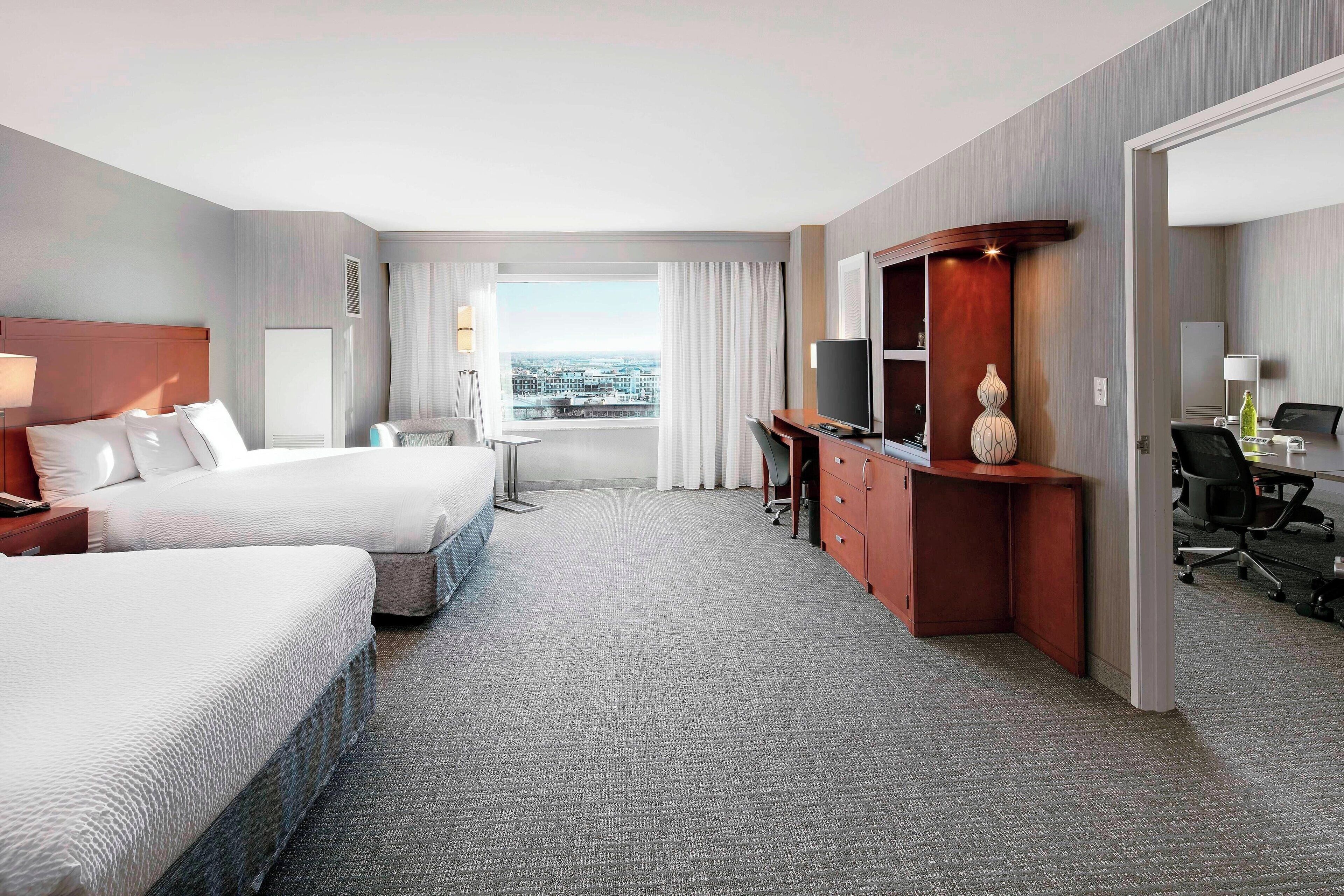 A hotel room with two beds, a desk, a TV, and a view of the city.