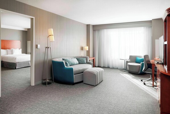 A modern hotel room with a grey sectional sofa, a single bed, a desk with a chair, and a large window with curtains.
