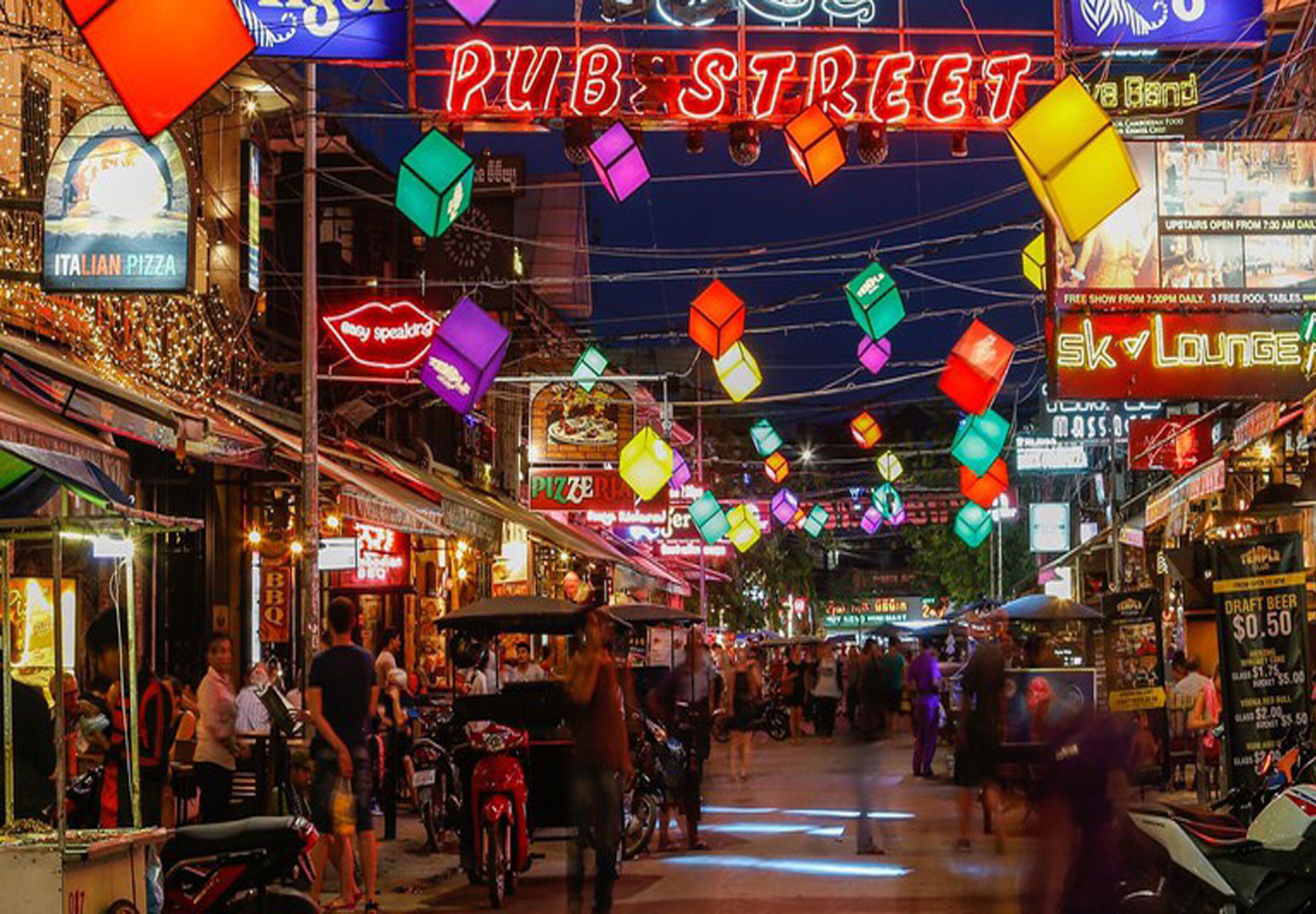 A busy street with colorful hanging decorations and various shops, including a pizza place and a sky lounge.