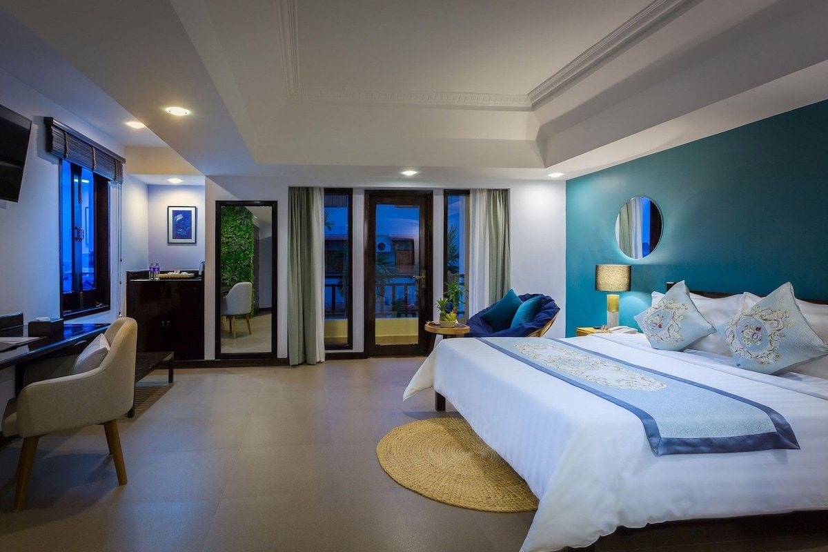Habitación de hotel con cama, escritorio, silla, televisor y balcón con cortinas.