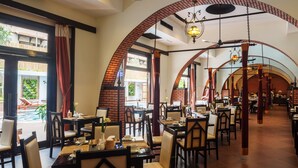 Um restaurante espaçoso com tetos altos, arcos de tijolo e lustres pendurados. As mesas estão dispostas com toalhas brancas e cadeiras, e áreas de estar ao ar livre são visíveis através de grandes janelas.