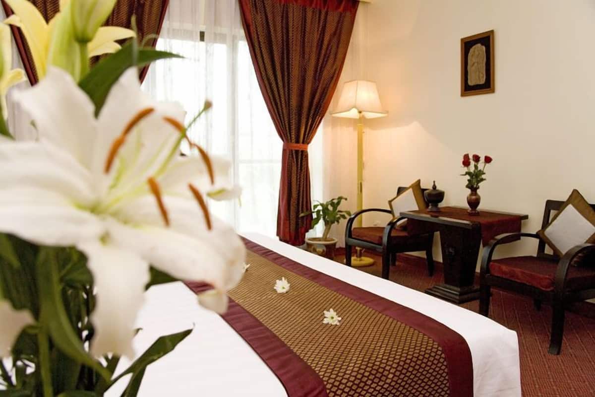 Una habitación de hotel con cama, escritorio, dos sillas, lámpara y un florero con flores.