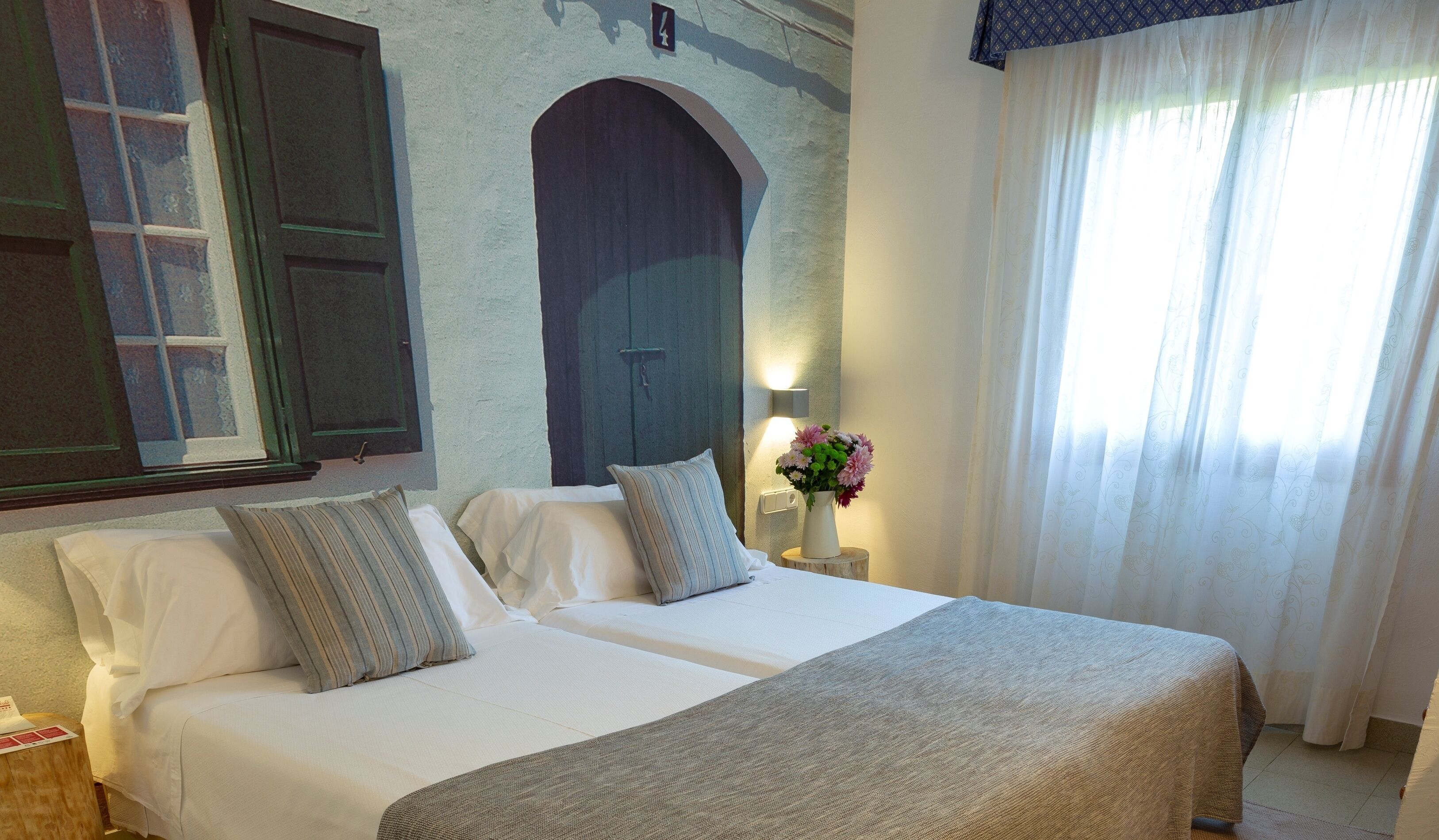 Habitación de hotel con dos camas, un amplio arco de entrada, una ventana con cortinas y una mesita de noche con un florero.