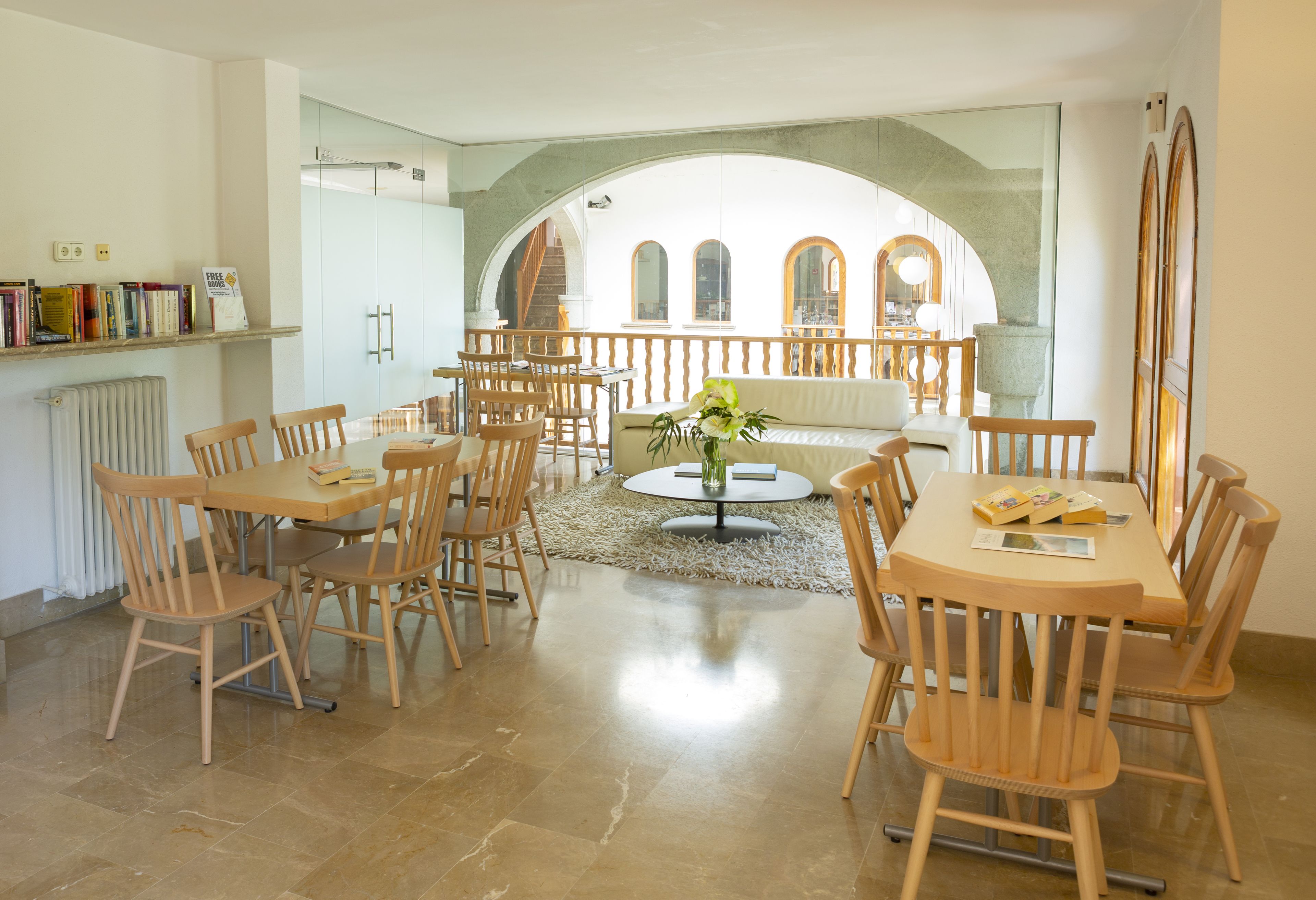 A spacious dining area with multiple wooden tables and chairs, a central round table with a vase, and a view through an arched window to a courtyard.
