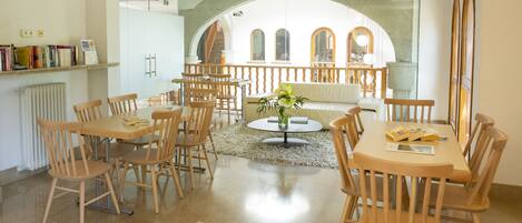 A spacious dining area with multiple wooden tables and chairs, a central round table with a vase, and a view through an arched window to a courtyard.