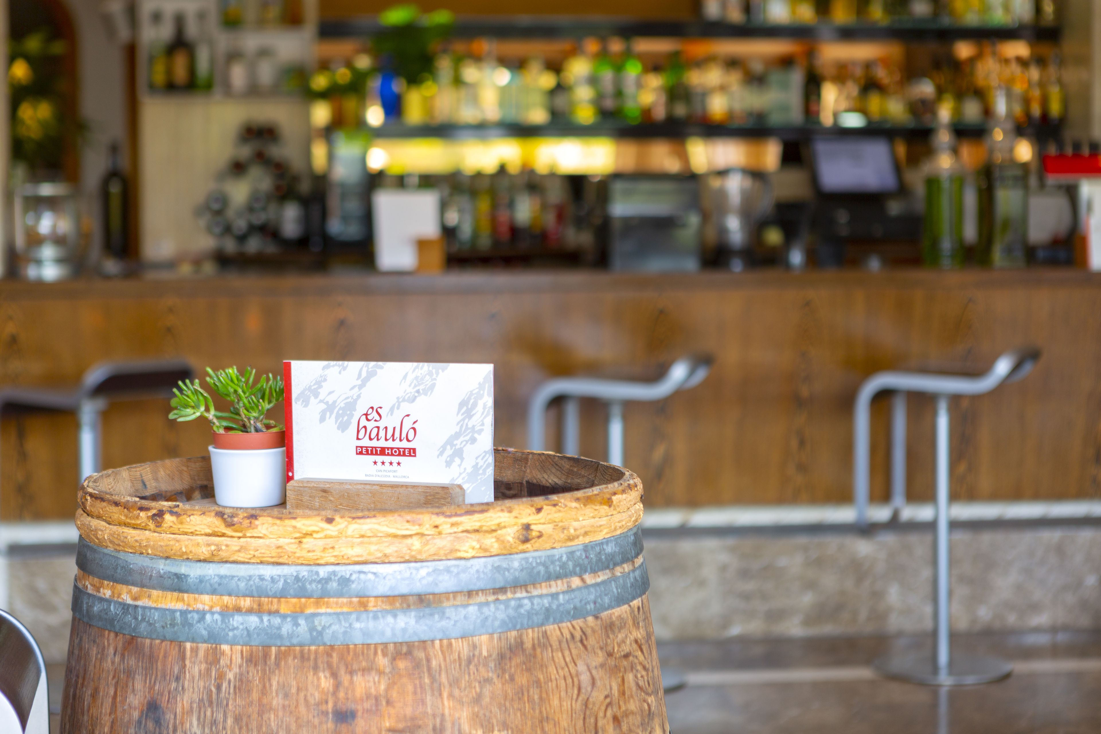 Un tonneau en bois surmonté d’une enseigne indiquant le « Baulo Private Hotel », une plante en pot et, au fond, un espace bar.
