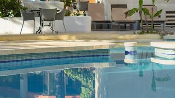 A clear swimming pool with a tiled edge, surrounded by lounge chairs and a table under a white umbrella.