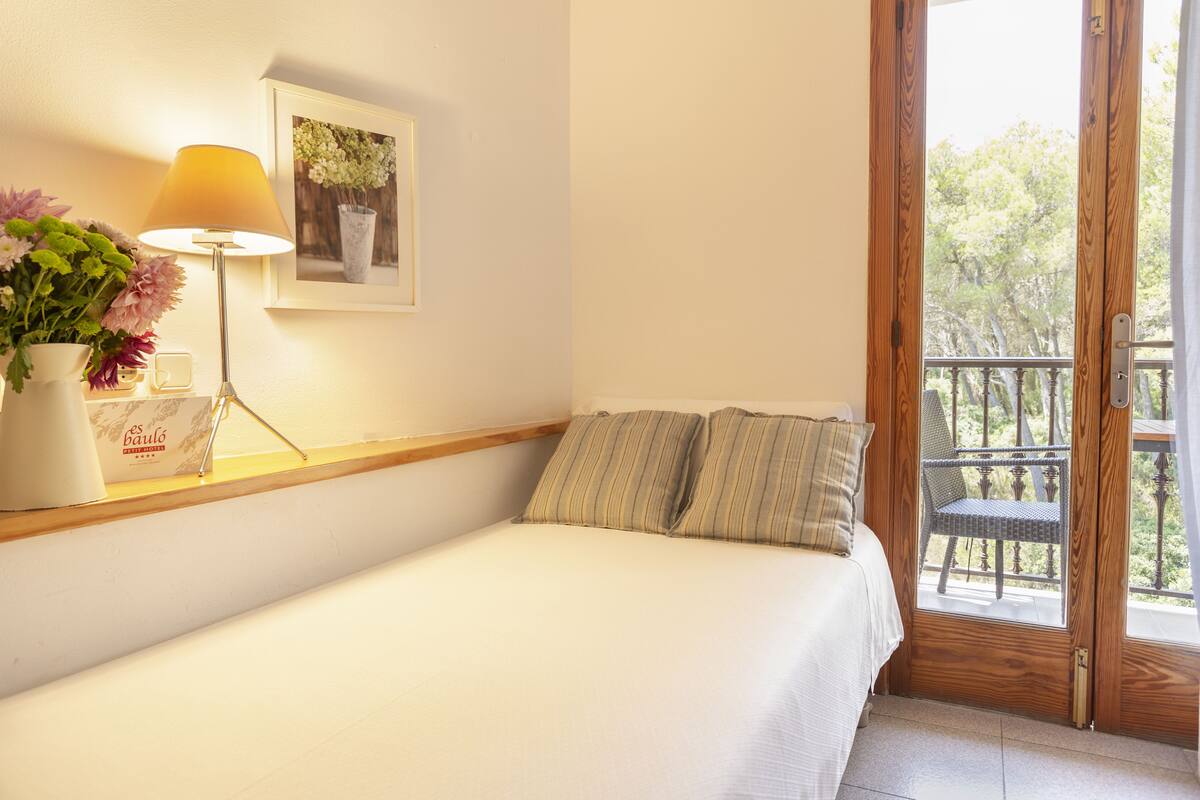 Un lit bien fait, avec une couette blanche, une lampe de chevet, un tableau encadré au mur et une fenêtre donnant sur des arbres.