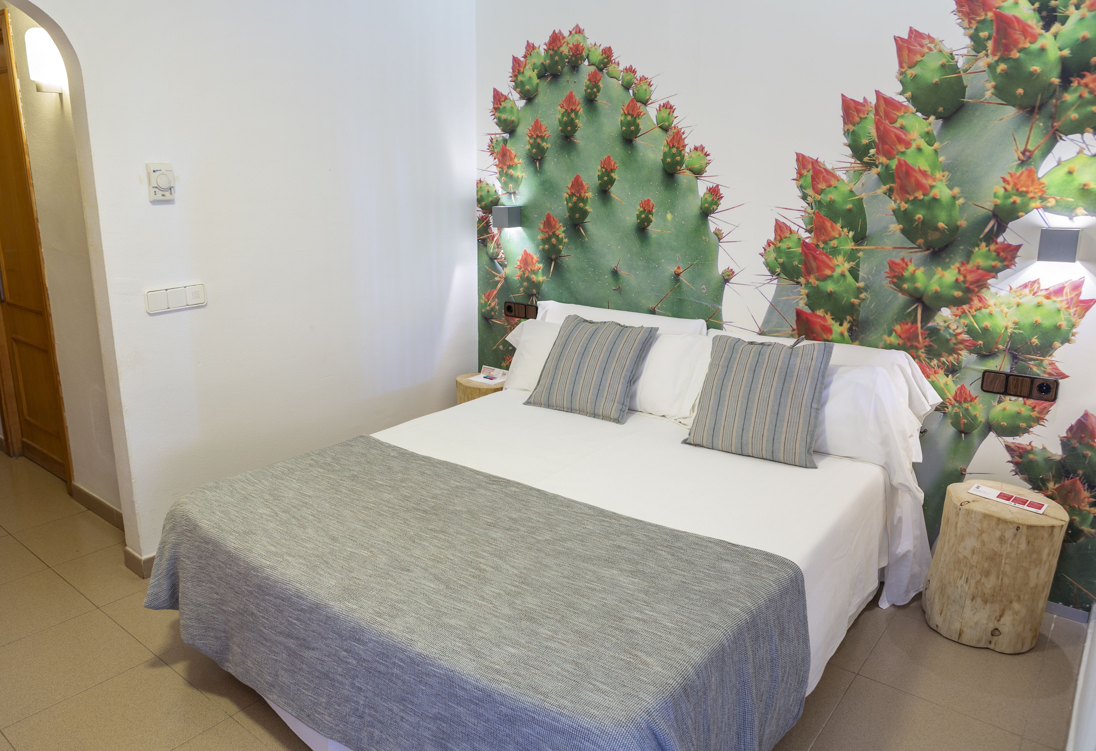 Ein Schlafzimmer mit Bett, Kissen, einem Nachttisch und Wandbild mit Kakteen.