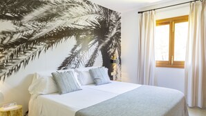 A bed with white bedding and grey pillows, a large wall mural of palm leaves, a wooden headboard, and a window with curtains.