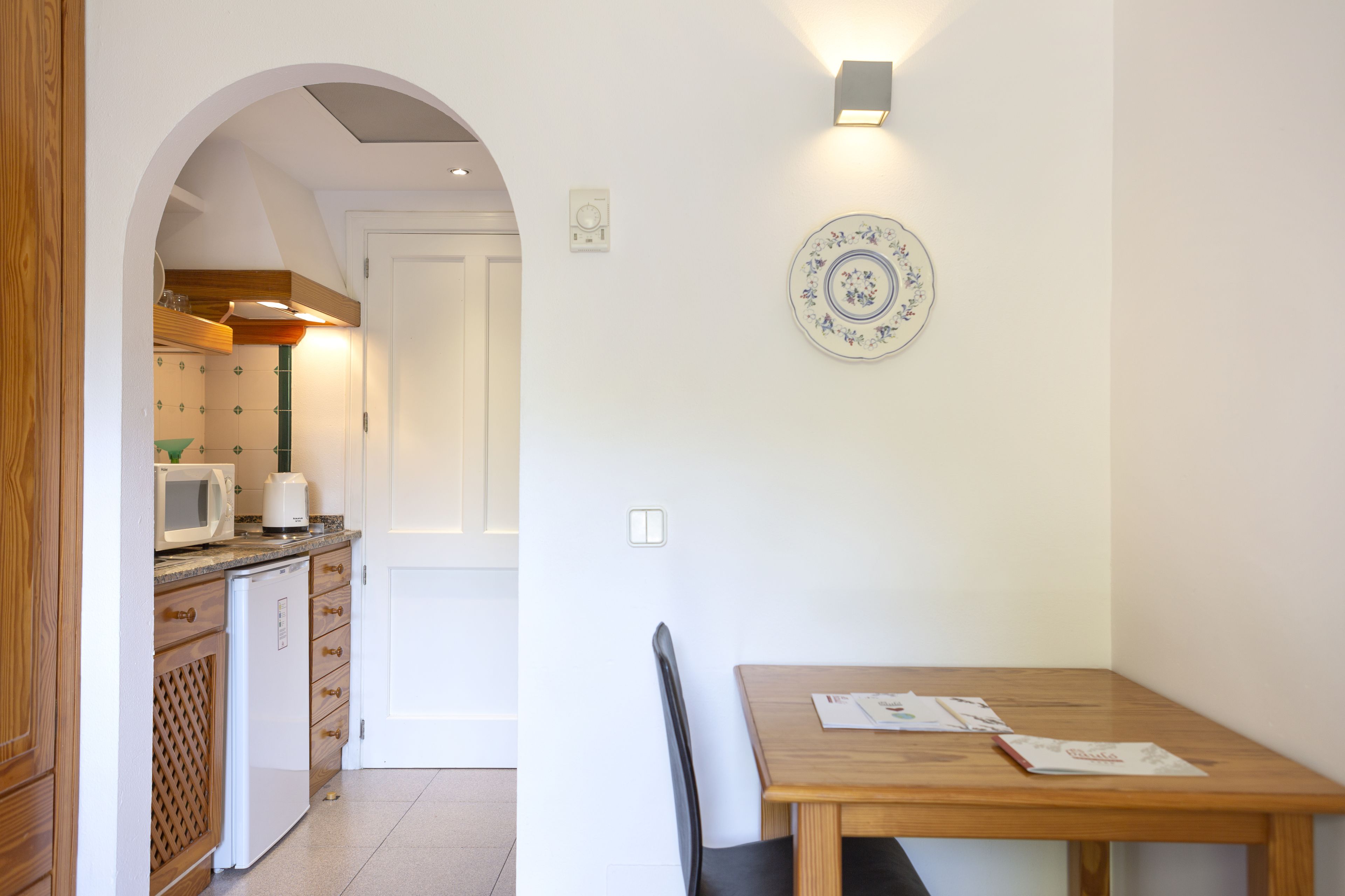 A kitchen with a microwave, toaster, and refrigerator, an archway leading to another room, and a dining table with chairs.