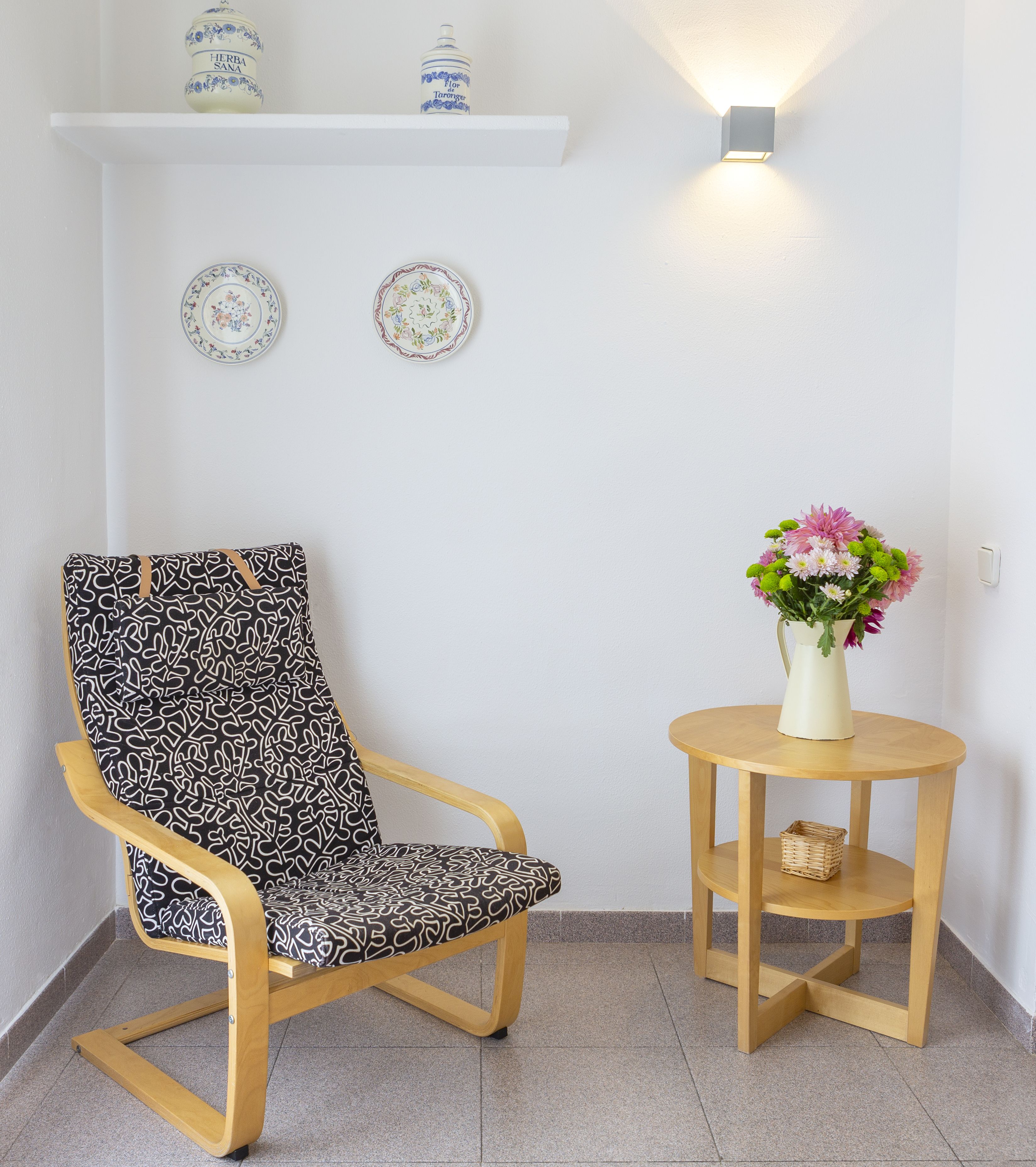 Una silla con un cojín estampado, una mesita redonda con un florero y dos platos decorativos en una estantería.