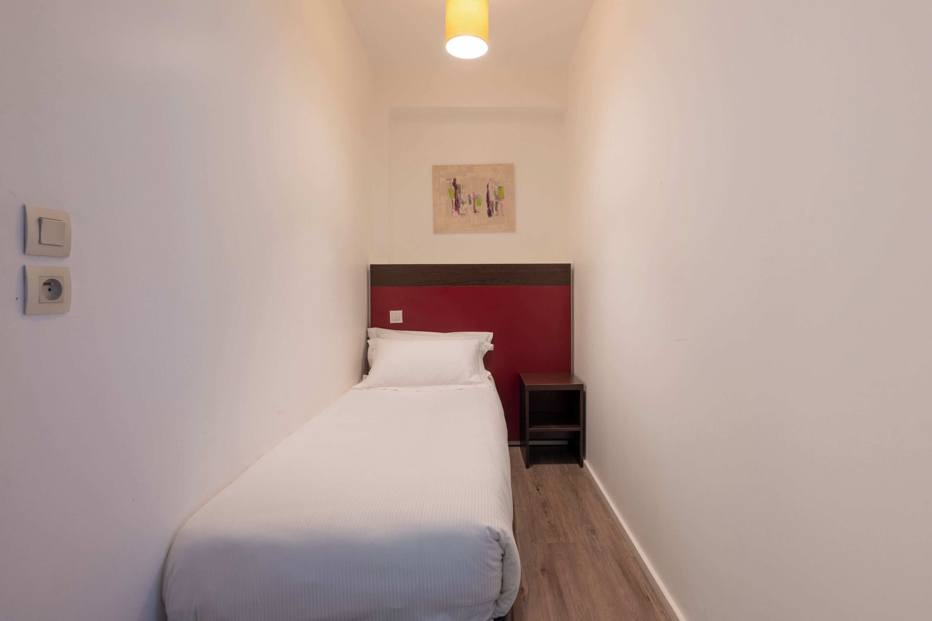 Une chambre d’hôtel étroite, avec un lit simple, une petite table de chevet et un luminaire fixé au mur.