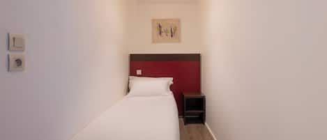 Une chambre d’hôtel étroite, avec un lit simple, une petite table de chevet et un luminaire fixé au mur.