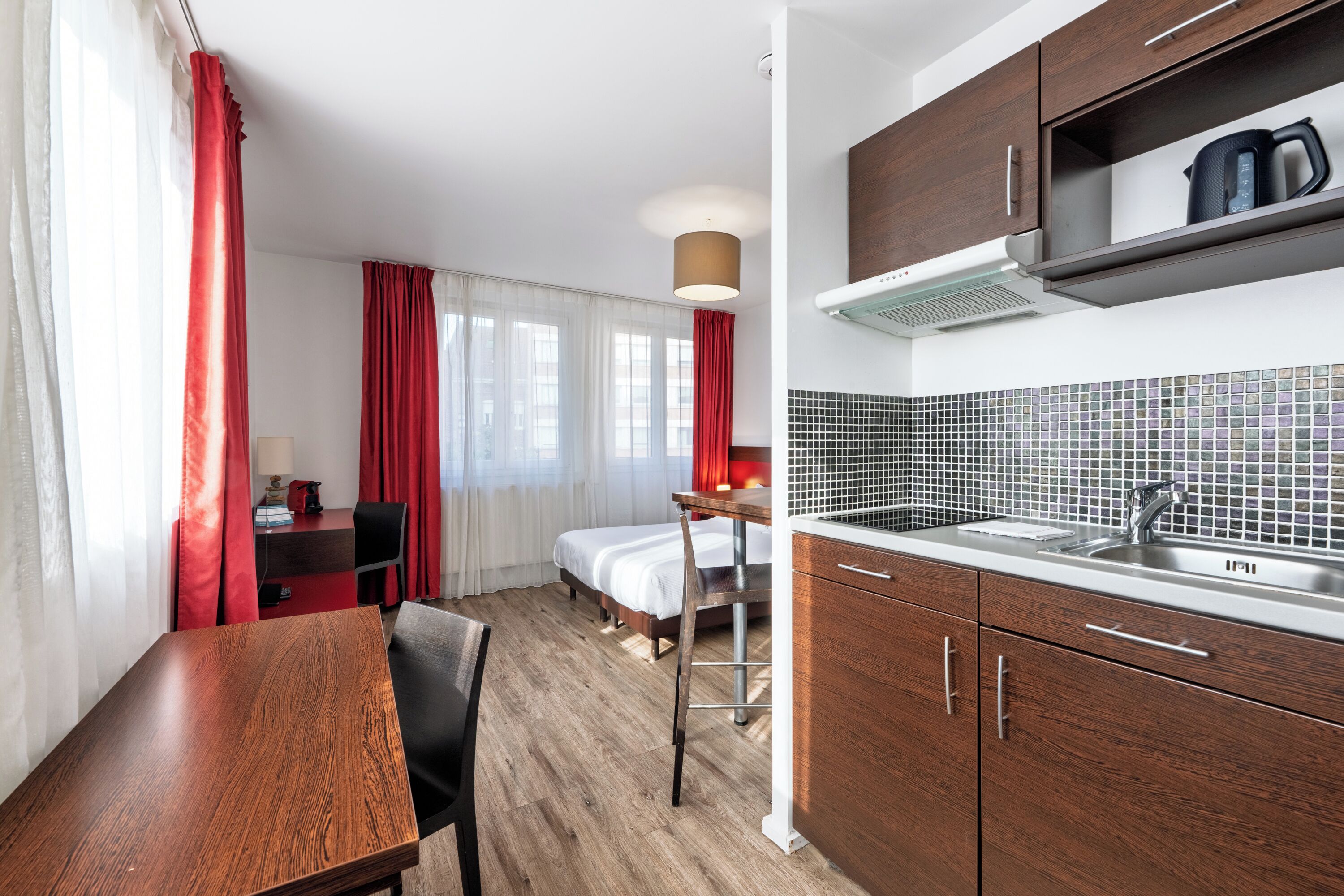 Una habitación de hotel moderna con una mesa de comedor de madera, una cama con ropa blanca, una kitchenette con un lavamanos de acero inoxidable y una cortina roja.