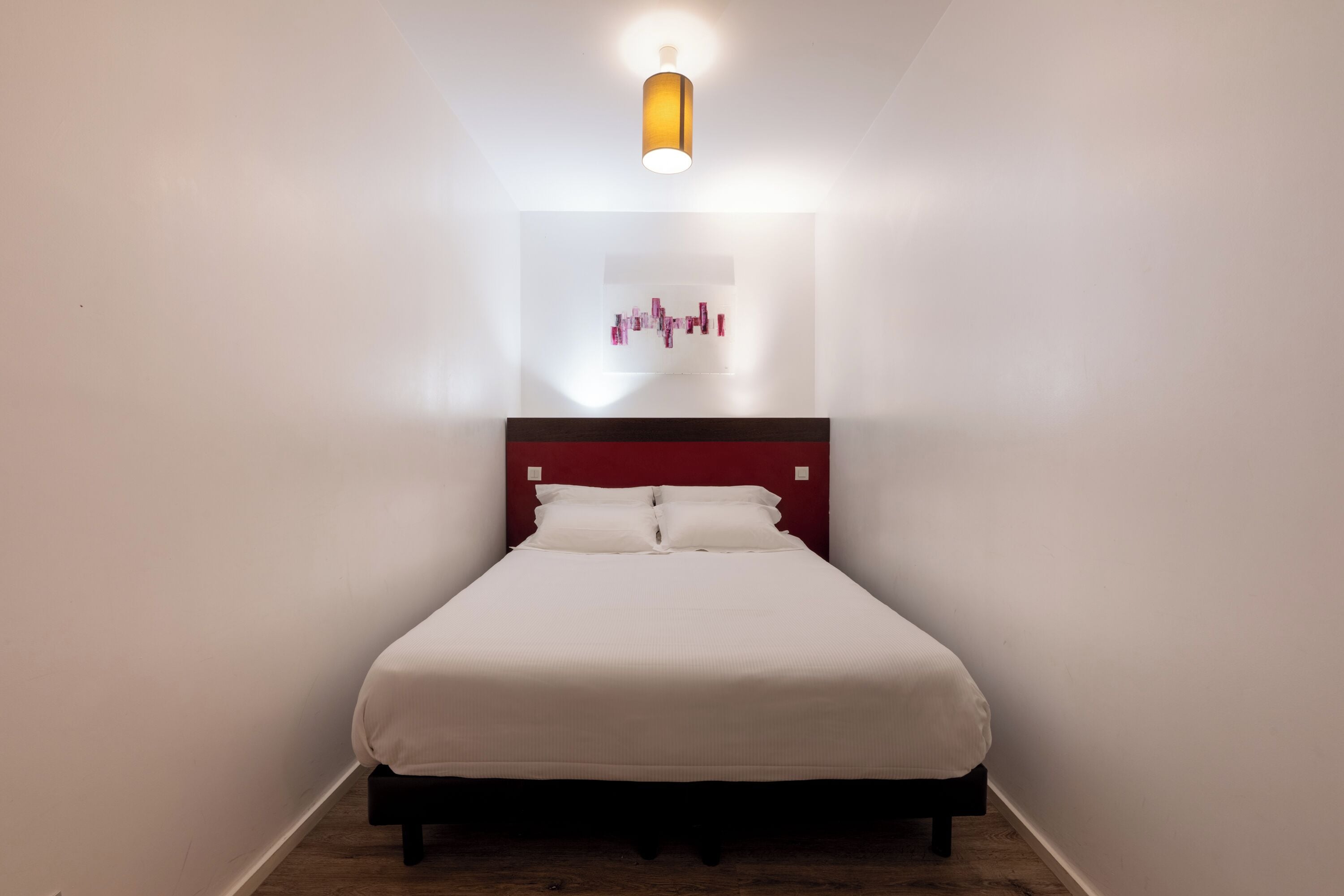 A neatly made bed in a room with white walls, a red headboard, and a modern light fixture above.