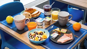 A breakfast tray with croissants, a bowl of cereal, a glass of orange juice, a mug, a bottle of jam, a carton of milk, a packet of yogurt, and a plate with ham and an orange.