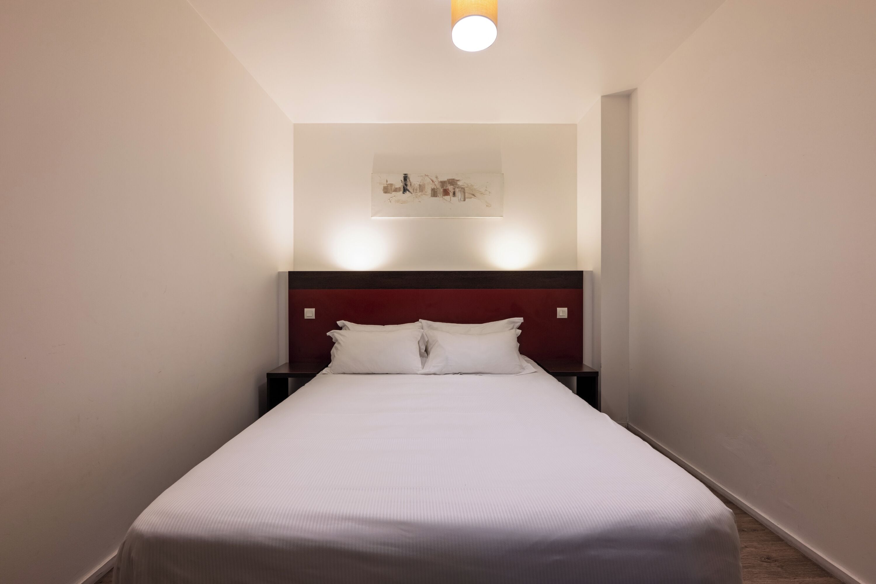 Un lit bien fait dans une chambre d’hôtel, avec une simple tête de lit et une seule œuvre d’art accrochée au mur.