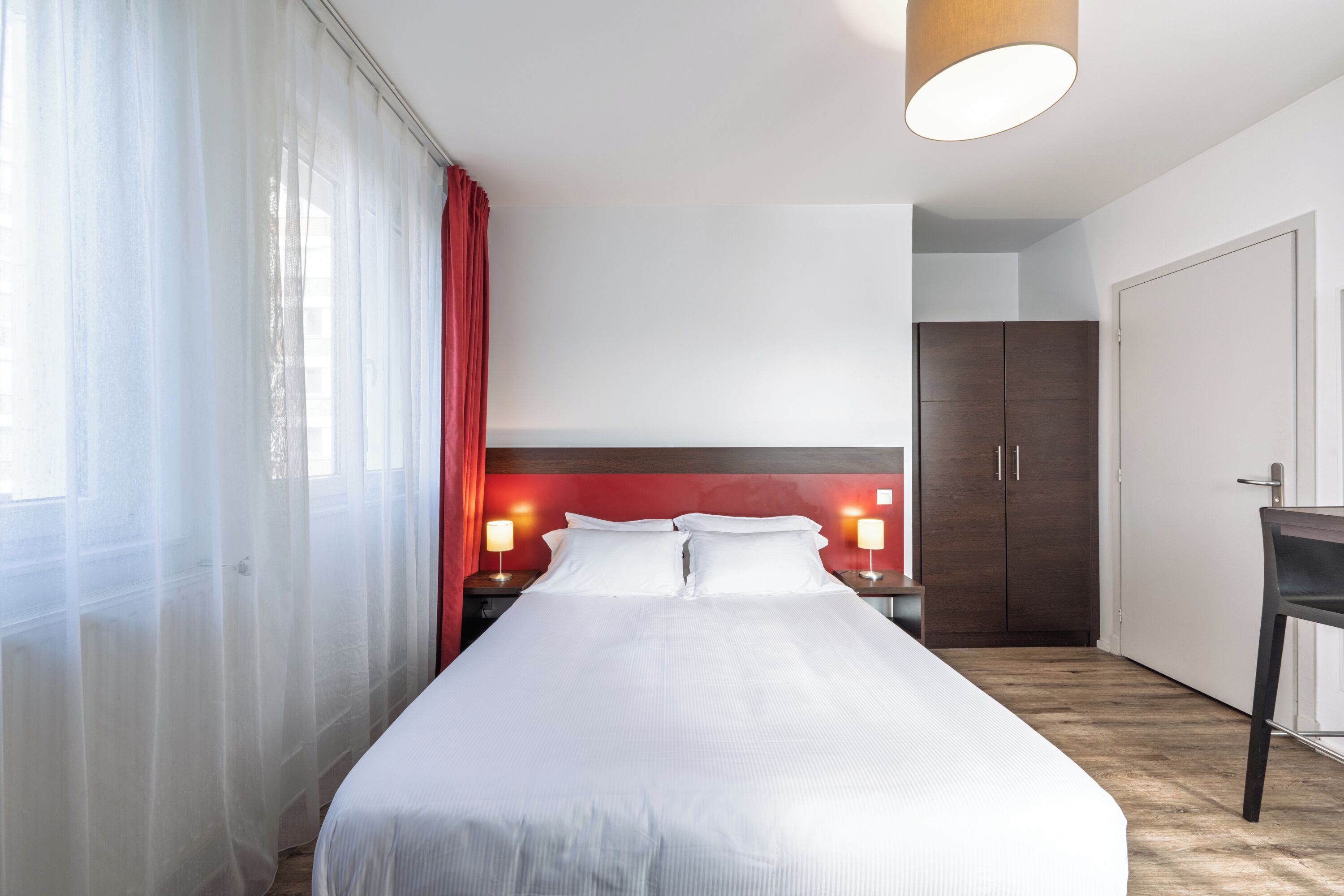 Hotellihuone, jossa on suuri sänky, puulattia, punainen verho ja työpöytä tuolilla.