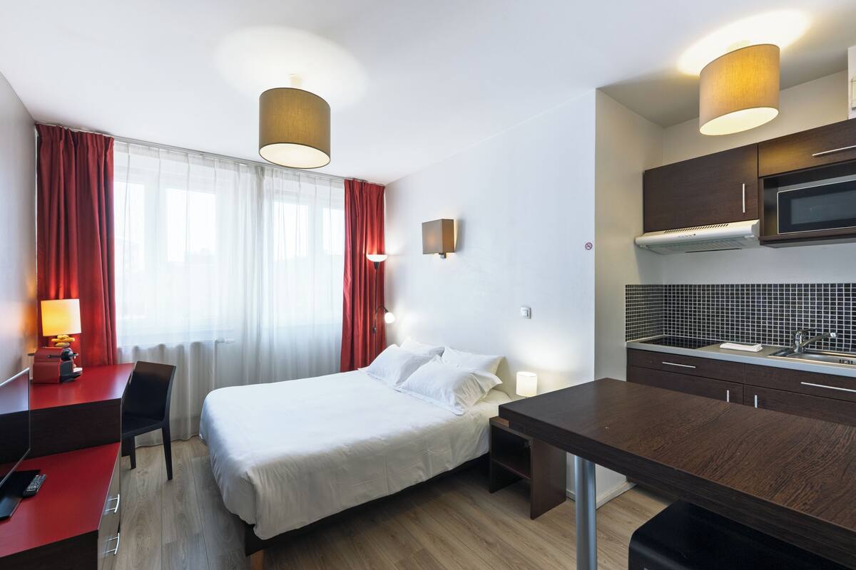 A hotel room with a bed, a desk, a chair, a small table, a microwave, and a sink.