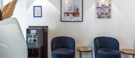 Un moderno vestíbulo de hotel con dos sillones azules, una pequeña mesa redonda, una caja fuerte y cuadros enmarcados en la pared.