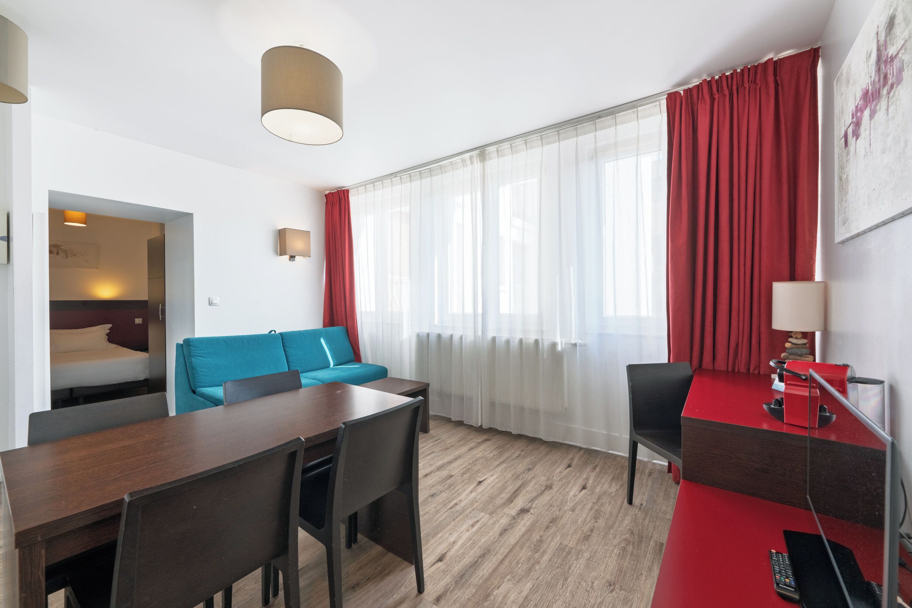 Habitación de hotel con piso de madera, una mesa de comedor con sillas, un sofá, un escritorio con lámpara y un ventanal con cortinas rojas.