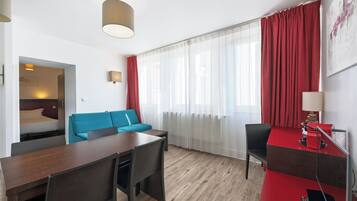 Habitación de hotel con piso de madera, una mesa de comedor con sillas, un sofá, un escritorio con lámpara y un ventanal con cortinas rojas.