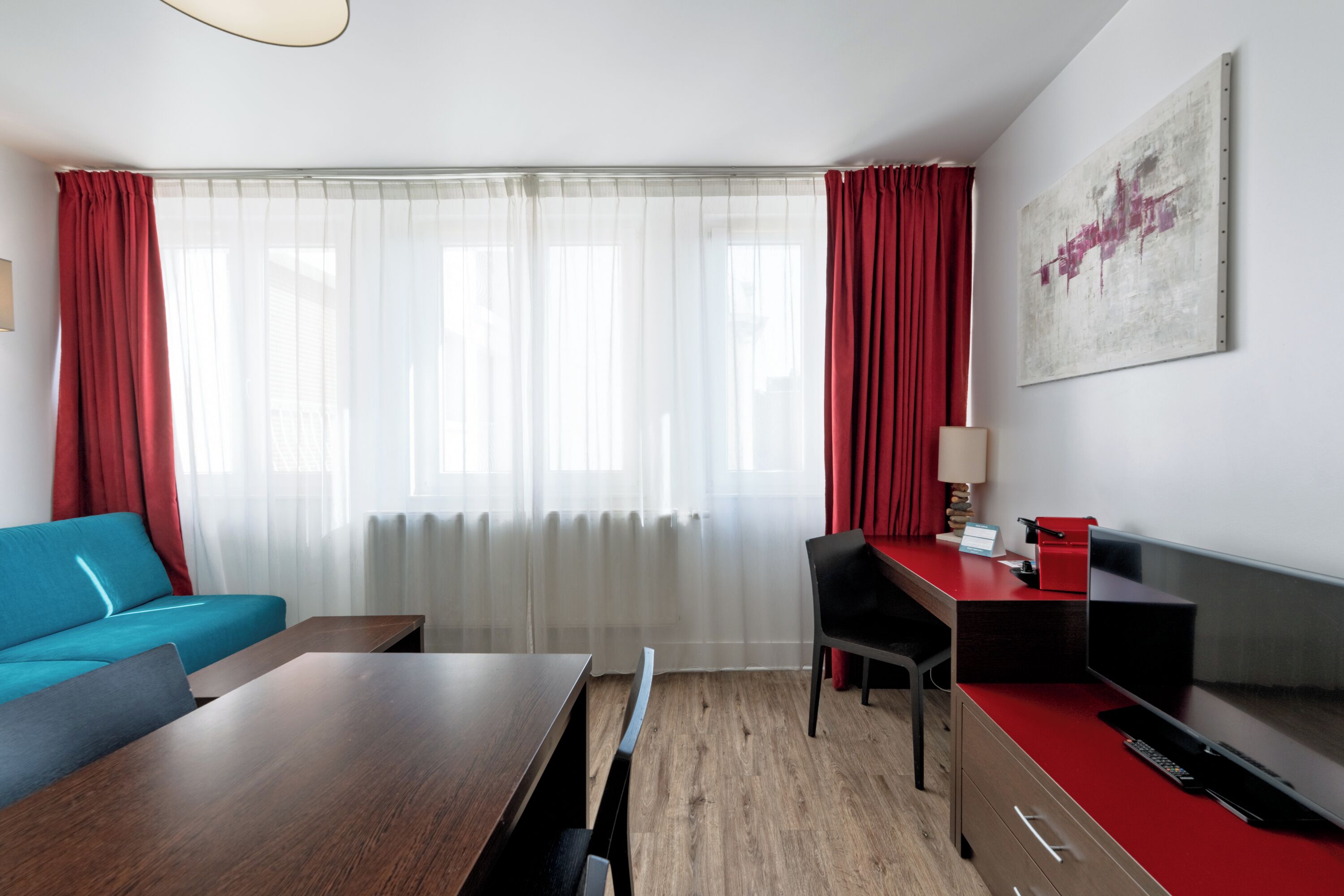 A modern hotel room with a wooden floor, a blue sofa, a wooden desk, a red chair, a TV, and a large window with sheer curtains.