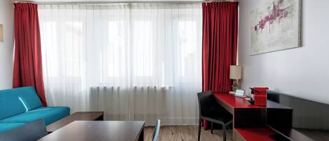 Una moderna camera d'albergo con pavimento in legno, un divano blu, una scrivania in legno, una sedia rossa, una TV e un'ampia finestra con tende trasparenti.