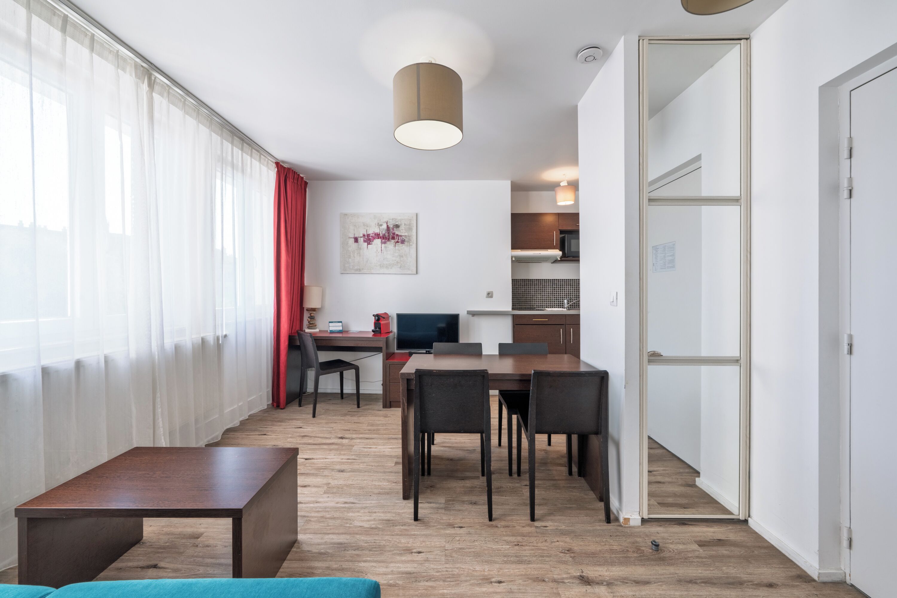 A modern living room with a wooden floor, a blue sofa, a dining table with chairs, a TV, and a kitchen area in the background.